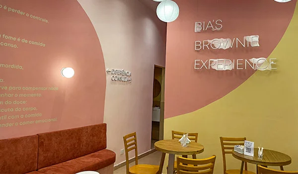 Foto da loja Bia´s Brownie Experience com paredes em tons de rosa e amarelo com mesas e cadeiras e sofa.