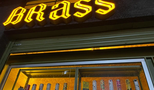 Estabelecimento com letreiro luminoso que diz 'BRASS', com destaque para a iluminação amarela, e exibe uma vitrine com produtos variados em uma loja de acessórios.