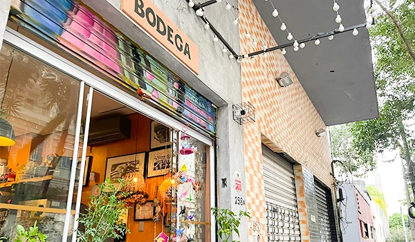 Fachada da bodega com abertura de vidro e decoração de interior visível, além de uma entrada com plantas ao redor e iluminação de cordão.