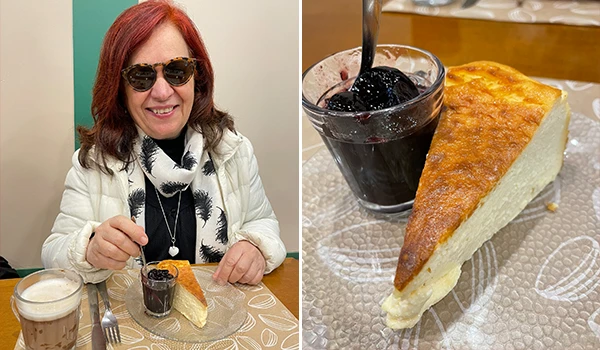 Deusa Rodrigues apreciando uma fatia de queijo cake com refrigerante escuro ao lado, em um ambiente de café ou padaria.
