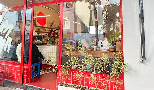 Foto da entrada do Drama Café com azulejos vermelhos e plantas em vasos do lado de fora e dentro clientes em espera na frente de balcão do estabelecimento.