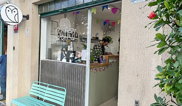Foto da frente da Gufi Café com balcão, vidro e na frente um simpático banco na cor azul.