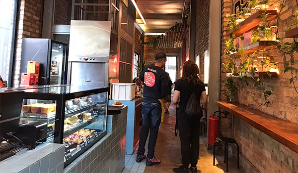 Interior de uma cafeteria moderna com clientes esperando na fila, paredes de tijolos expostos, iluminação aconchegante e decoração com plantas