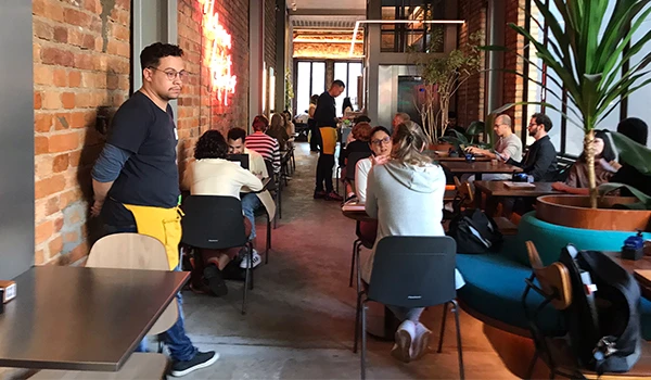 Ambiente aconchegante em um café ou restaurante movimentado com várias pessoas interagindo e um atendente em destaque ao lado, decor modernista com plantas e iluminação suspensa.