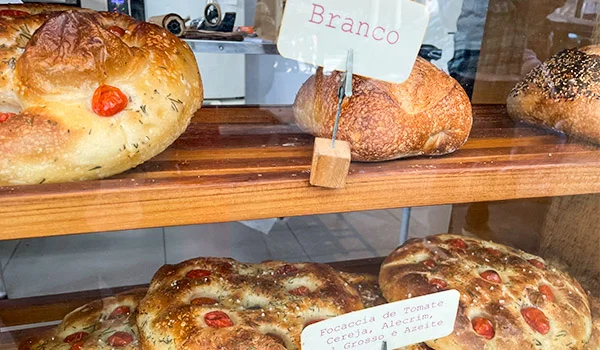Foto de uma vitrine com diversos pães e foccacia de tomate, cereja, alegrim e azeite.