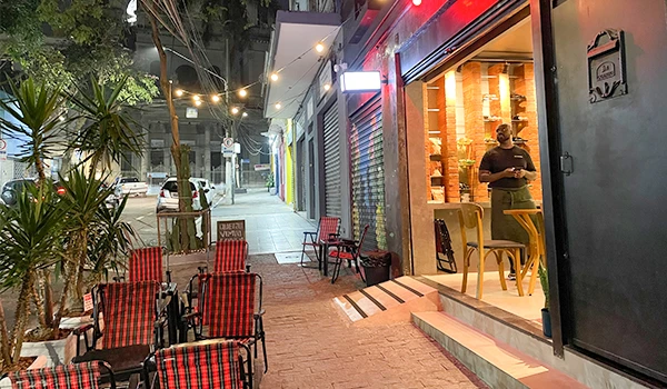 bar Tom Artesanal com mesas e cadeiras vermelhas e pretas em uma rua iluminada à noite em um bairro movimentado.