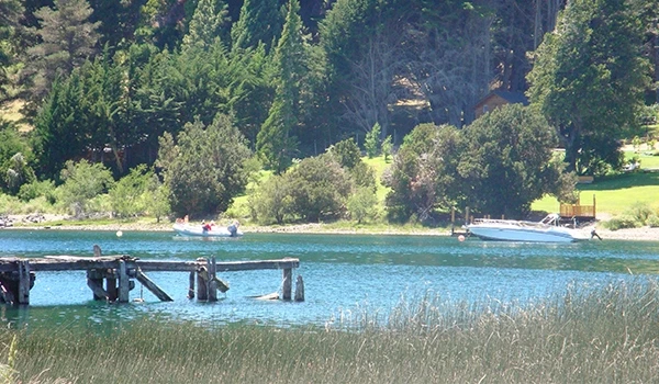 Linda paisagem natural com um lago tranquilo, cercado por árvores verdes e um pequeno cais de madeira. Barco ancorado e pessoas desfrutando da natureza.
