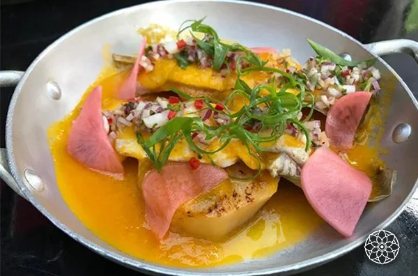Prato peruano servido em uma frigideira, com peixe coberto por molho amarelo, cebola roxa picada, ervas verdes e fatias de rabanete rosa.