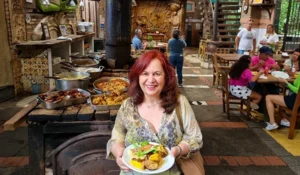 Mulher sorridente segurando um prato com comida típica em um restaurante decorado, enquanto as pessoas desfrutam do ambiente ao fundo.