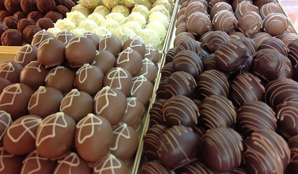 Vários tipos de trufas de chocolate decoradas com desenhos brancos e dourados, exibidas em uma vitrine de confeitaria, perfeitas para doces e festas.