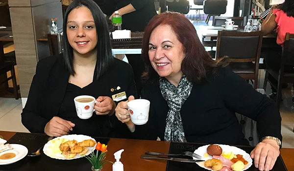 Deusa Rodrigues e funcionária do hotel desfrutam de um café da manhã com pratos típicos e bebidas quentes. Ambiente acolhedor e descontraído.