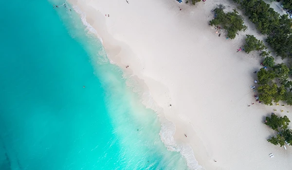 Vista aérea de uma praia paradisíaca com areia clara e águas cristalinas em tons de azul e verde, rodeada por vegetação.
