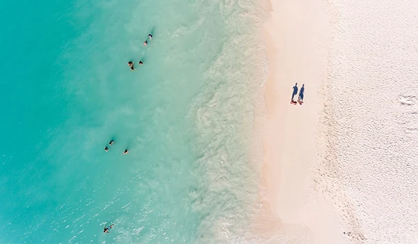 Vista aérea de uma praia com águas cristalinas e areia branca, onde pessoas estão nadando e um casal caminha pela orla, evocando momentos de verão.