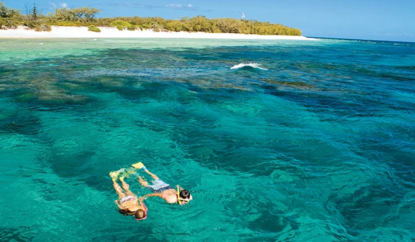 Pessoa nadando em águas cristalinas na praia, com areia branca ao fundo e céu claro azul, ideal para quem busca relaxar e aproveitar o mar tropical. Rota de Viagem para a Oceania.