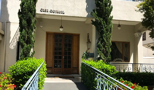 Entrada do prédio com varanda, jardim bem cuidado, árvores e plantas verdes, acesso com degraus, fachada branca, ambiente externo iluminado, ideal para morar ou investir