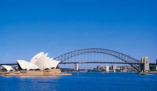 Vista da Sydney Opera House e da Harbour Bridge ao longo da baía de Sydney em um dia ensolarado, com céu azul e água tranquila. Rota de Viagem para a Oceania.