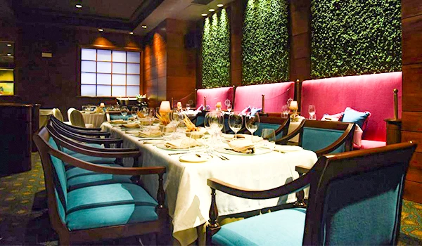 Salão de restaurante elegante com mesas arrumadas para jantar, cadeiras azuis e decoração moderna com paredes verdes e iluminação aconchegante.