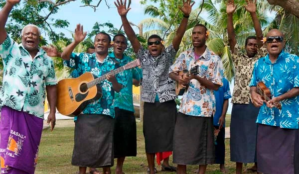 Grupo de homens sorrindo, cantando e tocando violão ao ar livre, em uma celebração cultural em um ambiente tropical com árvores e céus azuis ao fundo.