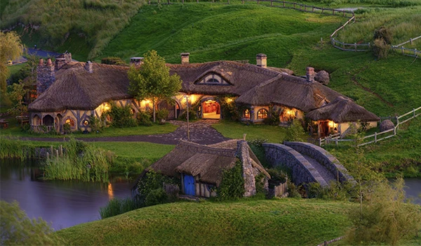 Imagem de uma casa de campo artesanal com telhados de barro em uma paisagem verdejante, com lago e vegetação ao redor, iluminada à noite, criando uma atmosfera acolhedora e rústica. Nova Zelândia - Rota de Viagem para a Oceania.