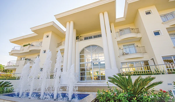 Imagem de um hotel luxuoso com fachada branca, janelas grandes, varandas e uma fonte na frente, com jardins bem cuidados.