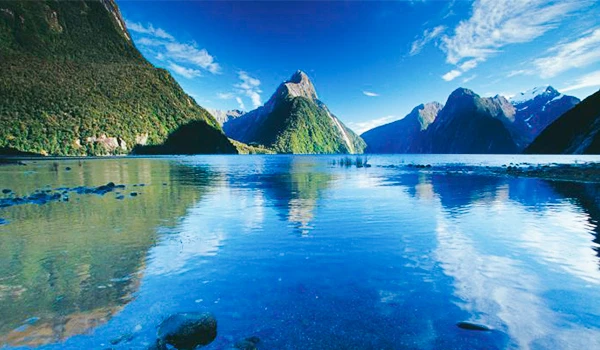 Paisagem de fiorde com montanhas verdes e águas cristalinas refletindo o céu azul claro, capturando a beleza natural de uma região de natureza exuberante. Nova Zelândia - Rota de Viagem para a Oceania.