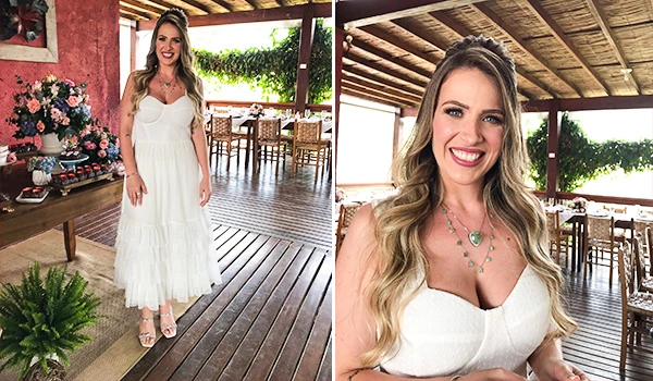 Mulher sorridente com vestido branco elegante em ambiente rústico ao ar livre, decoração com flores e móveis de madeira, cenário ideal para fotos especiais.