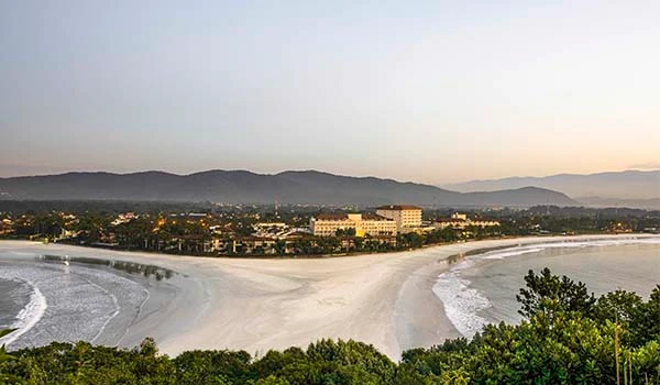 Vista panorâmica da praia do Guarujá com mar, areia, vegetação e montanhas ao fundo ao pôr do sol, em uma tarde de céu limpo e atmosfera tranquila.