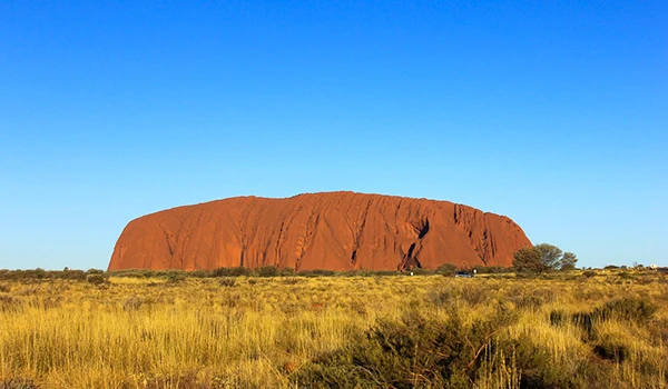 Imagem da formação rochosa Uluru na Austrália, destacada pelo céu azul claro e o cenário de campo aberto ao seu redor. Rota de Viagem para a Oceania.