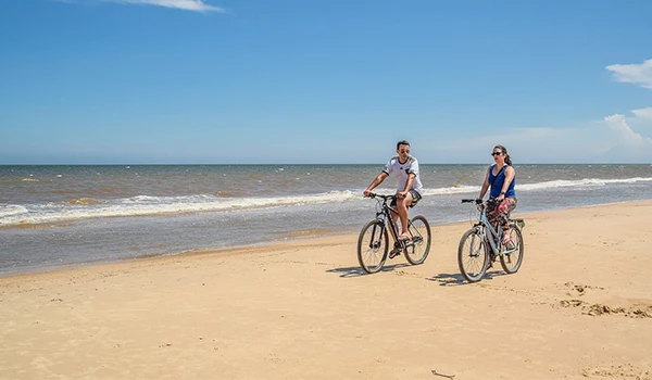 Homem e mulhere curtindo passeio de bicicleta na praia ensolarada com céu azul e mar ao fundo, aproveitando o dia de lazer.