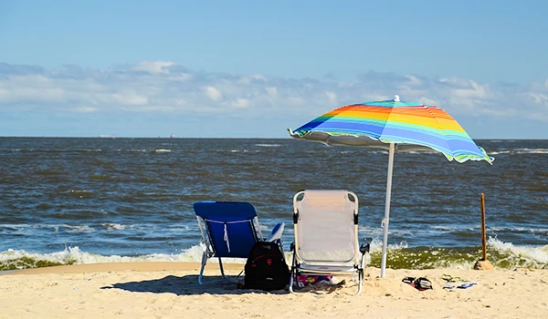 Praia com cadeiras, guarda-sol colorido e mochila na areia, cenário de lazer ao ar livre, ideal para relaxar e curtir o mar em um dia ensolarado.
