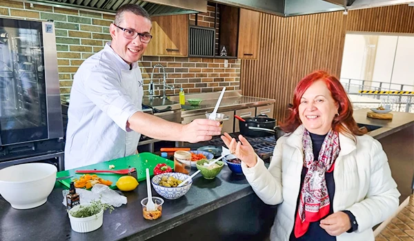 Foto do Chef Cassio Prados e Deusa Rodrigues na cozinha do Espaço Gastronômico do Mercadão, onde ele preparou uma oficina, um ambiente moderno com bancada de mármore e utensílios de cozinha ao redor.