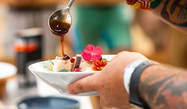 Prato de comida com molhos e flores decorativas sendo servido por uma pessoa com tatuagens na mão, cenário de cozinha ao fundo.