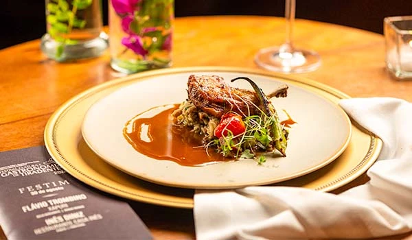 Prato sofisticado de gastronomia com carne e acompanhamentos no centro de uma mesa de jantar elegante, com taças e copos ao fundo.