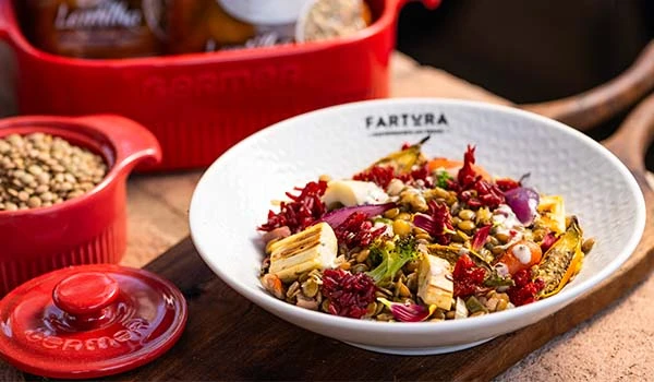 Salada de legumes e grãos colorida em uma tigela branca, com ingredientes frescos como beterraba, milho, tomate e alface, sobre uma mesa de madeira.