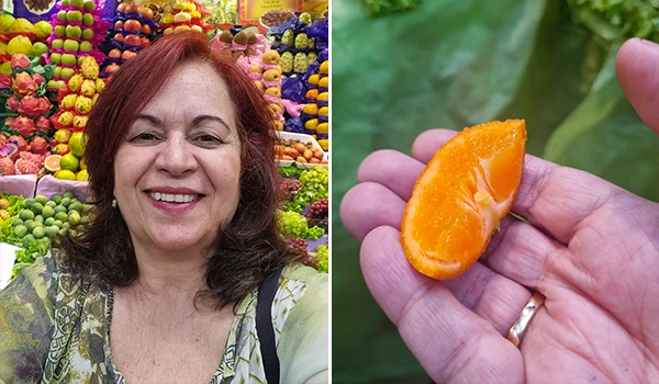 Deusa Rodrigues segurando uma fatia de laranja, com fundo de feira de frutas coloridas incluindo maçãs, bananas e morangos, destacando a variedade de frutas frescas. Onde comer no Mercadão.