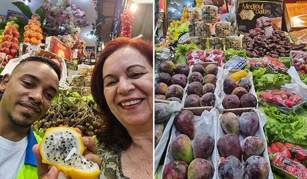 Imagem de uma barrada de frutas variadas, incluindo mamão, goiaba, romã, e outras, com duas pessoas sorrindo ao fundo, uma delas segurando uma fruta cortada, no Mercadão de SP.