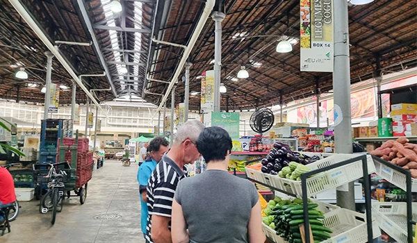 Pessoas fazem compras em mercado com destaque para frutas e verduras expostas na seção de hortifrúti, sob teto de madeira.