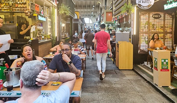 Imagem de uma rua de comércio movimentada com várias pessoas desfrutando de momentos sociais, lojas e restaurantes ao longo da calçada, promovendo a experiência de compras e lazer ao ar livre. Onde comer no Mercadão.