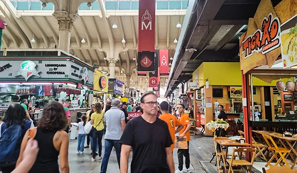 Imagem de um movimentado mercado interno com várias lojas, pessoas caminhando, algumas de óculos, em um ambiente com alto teto e iluminação, no Mercadão de SP.