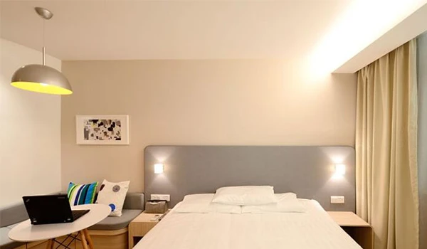 Imagem de um quarto de hotel moderno com cama de casal, cabeceira iluminada, mesa com laptop e uma poltrona ao lado, decorado com cortinas claras e quadro na parede.