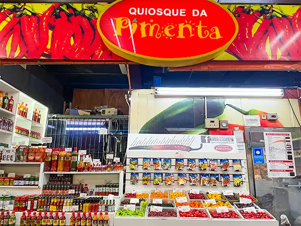 Quiosque da Pimente com frutas, temperos e produtos alimentícios variados, localizado em uma feira ou loja, com destaque para a sinalização 'Quiosque da Pimente'.