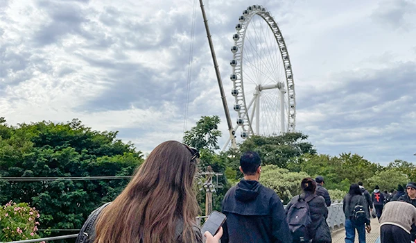 Turistas aproveitando o passeio na cidade, com uma roda gigante ao fundo e céu parcialmente nublado