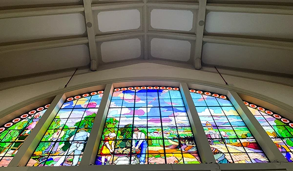 Vitral colorido em uma igreja, mostrando pessoas em diferentes atividades ao ar livre, com árvores e céu ao fundo, sob uma estrutura de teto branca.