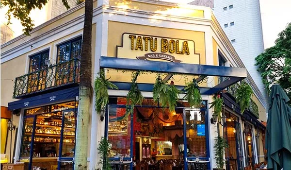 Fachada de um restaurante chamado “Tatu Bola Bar & Grill”, em um edifício de estilo clássico, com paredes em tons de bege e branco. A entrada tem grandes janelas de vidro, toldos escuros e vasos com plantas pendentes. Mesas e cadeiras ocupam a área externa, enquanto o interior iluminado exibe garrafas e decoração. Árvores e um prédio alto aparecem ao fundo.