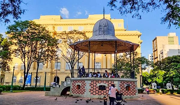 Praça arborizada com um coreto ornamentado ao centro, em frente a um edifício histórico amarelo. Há pessoas caminhando e sentadas ao redor, além de árvores, bancos e algumas aves no chão, sob um céu azul.