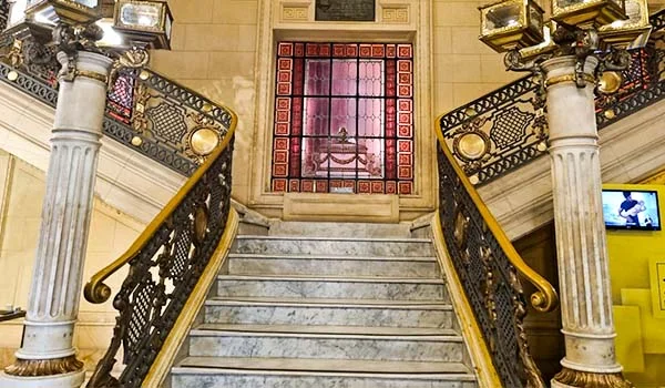Escadaria interna de um edifício histórico, com degraus de mármore, corrimãos ornamentados em ferro e dourado, colunas de mármore branco e uma janela decorativa ao fundo.
