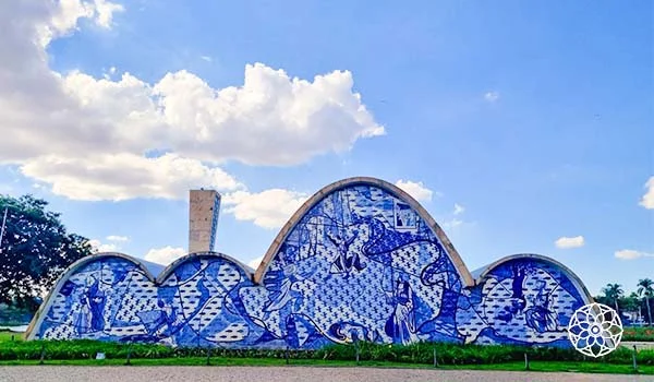 Construção de arquitetura modernista com três grandes arcos revestidos por painéis de azulejos azuis e brancos, decorados com figuras humanas, animais e padrões geométricos. Ao redor há gramado e árvores, sob um céu azul com grandes nuvens brancas.