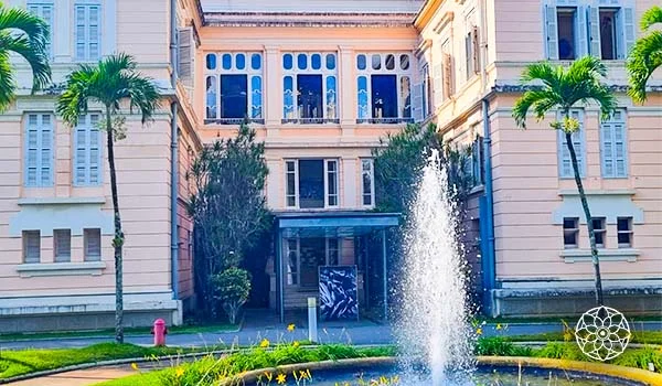 Visita ao Palácio da Liberdade. Fachada de um palácio histórico em estilo clássico, com paredes em tons claros, janelas altas com venezianas e detalhes ornamentais. Ao centro, há uma entrada com cobertura de vidro, cercada por árvores e jardins. Em primeiro plano, uma fonte circular lança um jato de água para o alto, com palmeiras nas laterais.
