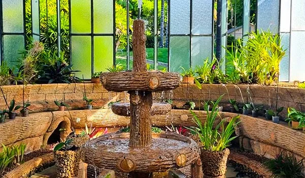 Fonte de água ornamental de madeira, com três níveis circulares, em um jardim interno repleto de plantas tropicais e vasos, diante de grandes janelas de vidro.