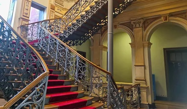 Escadaria interna ornamentada, com degraus revestidos de carpete vermelho e corrimãos de madeira. As laterais apresentam elaborados gradis de ferro trabalhado com arabescos. A escada se divide em diferentes lances junto a um amplo espaço interno decorado com arcos, colunas e detalhes arquitetônicos clássicos em tons de bege, verde e dourado.